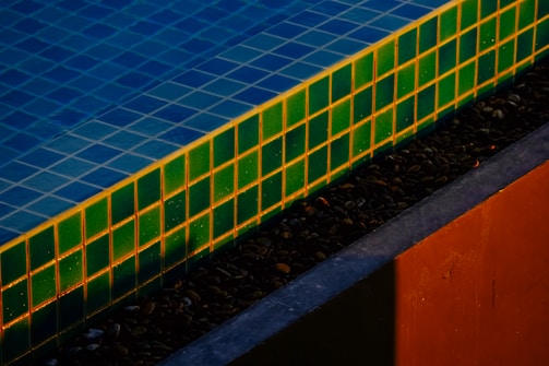Colorful tiles in green and blue form the edge of a swimming pool, with sunlight creating distinct shadows and highlights. The texture of small pebbles is visible beside the pool.