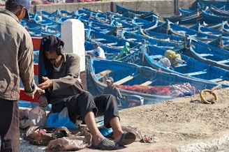 A man is sitting on a concrete surface surrounded by plastic bags, appearing to exchange something with another person. In the background, numerous blue wooden boats are docked tightly together, suggesting a busy fishing harbor. Various fishermen can be seen attending to their boats or resting.