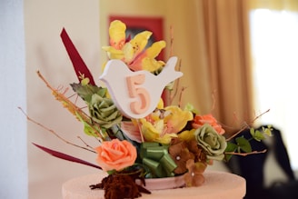 A decorative floral arrangement features a mix of colorful artificial flowers, including yellow orchids, orange and green roses, and brown blooms. A prominent white bird-shaped card with the number 5 is placed centrally. The composition includes green leaves and branches as accents, all set on a neutral-toned background.