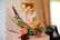 A decorative floral arrangement features a mix of colorful artificial flowers, including yellow orchids, orange and green roses, and brown blooms. A prominent white bird-shaped card with the number 5 is placed centrally. The composition includes green leaves and branches as accents, all set on a neutral-toned background.