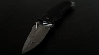 A close-up view of a folding knife with a partially serrated blade lying on a dark surface. The blade has water droplets on it, and the handle appears to be textured for grip.