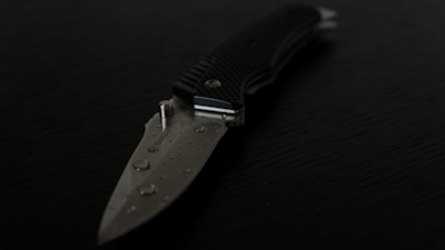 A close-up view of a folding knife with a partially serrated blade lying on a dark surface. The blade has water droplets on it, and the handle appears to be textured for grip.