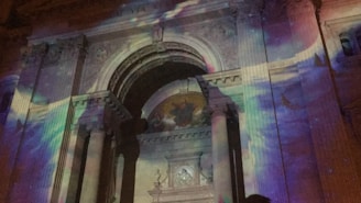 The facade of a classical building with ornate columns and an arch is illuminated by colorful lights, creating a vibrant display. The projected lights feature swirling patterns in hues of purple, blue, and pink, adding a dynamic and artistic touch to the architectural details.