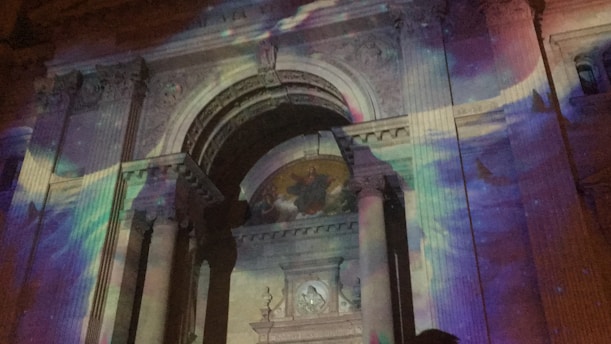 The facade of a classical building with ornate columns and an arch is illuminated by colorful lights, creating a vibrant display. The projected lights feature swirling patterns in hues of purple, blue, and pink, adding a dynamic and artistic touch to the architectural details.