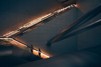 A modern architectural setting featuring an illuminated escalator with two people ascending. The scene includes sleek metallic beams and dimly lit walls, creating a futuristic and urban ambiance.