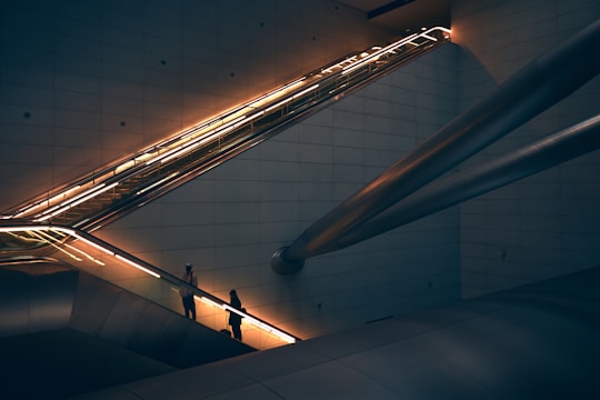A modern architectural setting featuring an illuminated escalator with two people ascending. The scene includes sleek metallic beams and dimly lit walls, creating a futuristic and urban ambiance.