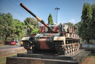 A large military tank is displayed on a raised platform outdoors, surrounded by trees and against a blue sky. The tank is painted in camouflage colors with a long barrel pointing slightly upward. In the background, there is a road with a few vehicles and power lines. A plaque on the platform appears to display information about the tank.
