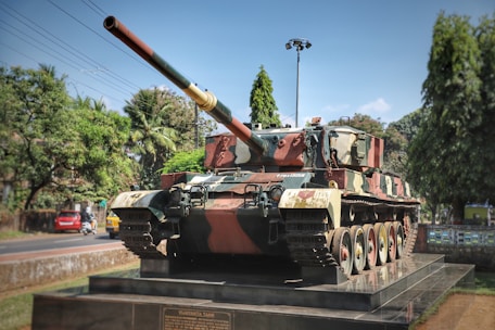 A large military tank is displayed on a raised platform outdoors, surrounded by trees and against a blue sky. The tank is painted in camouflage colors with a long barrel pointing slightly upward. In the background, there is a road with a few vehicles and power lines. A plaque on the platform appears to display information about the tank.