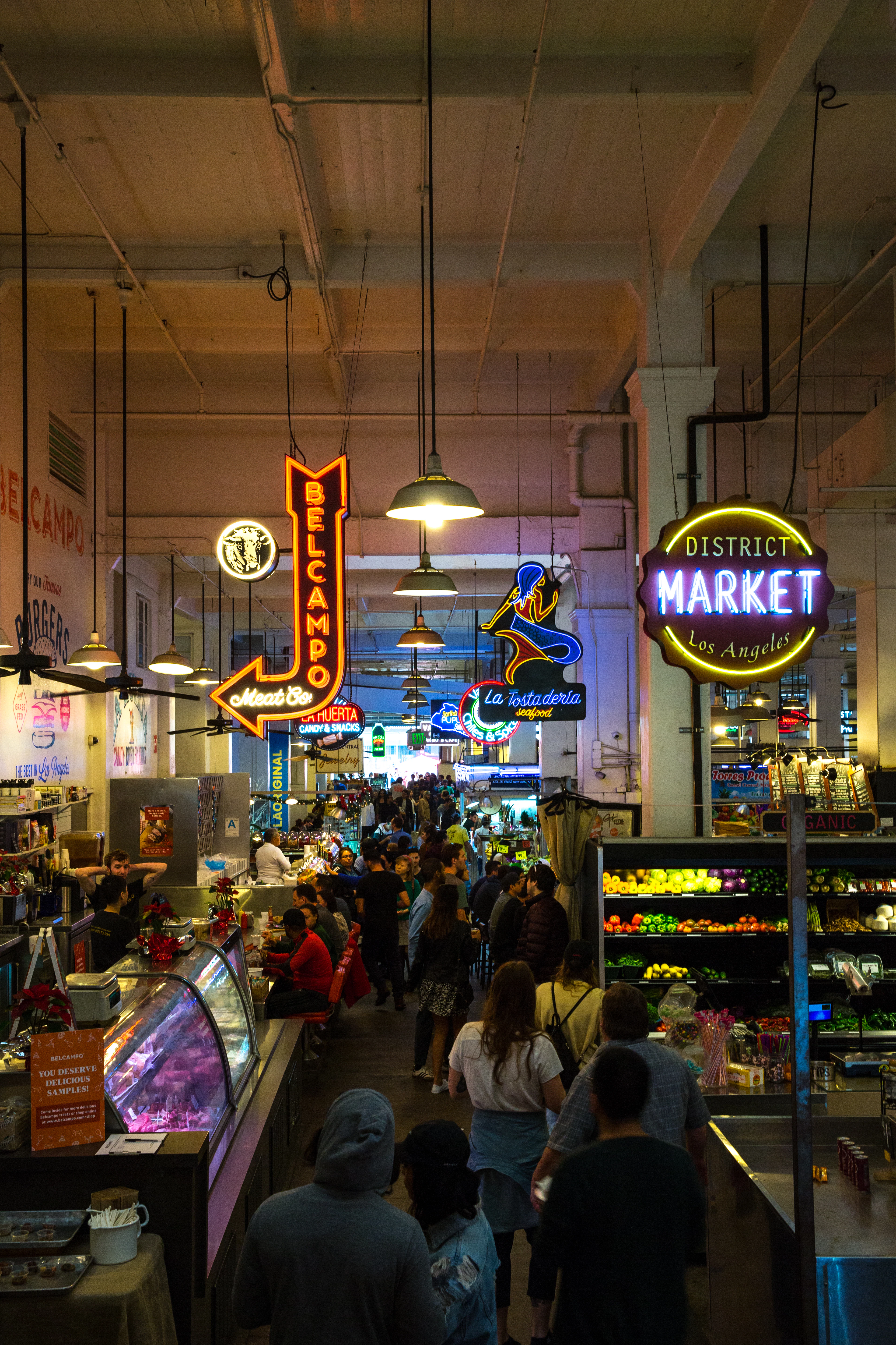 Neon stall signs glowing under the high ceiling of Grand Central Market in downtown Los Angeles, shoppers moving down the central aisle between a butcher's display case and racks of bright produce.