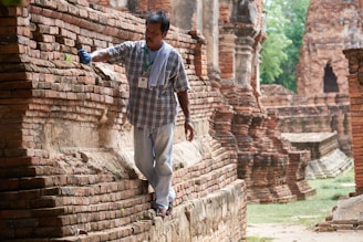 A man wearing a plaid shirt and jeans carefully brushes the surface of ancient brick ruins. He is holding a brush in one hand and has a towel draped over his shoulder. The setting appears to be an archeological site or historical monument with weathered brick structures in the background.