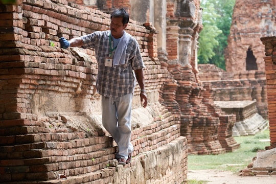 A man wearing a plaid shirt and jeans carefully brushes the surface of ancient brick ruins. He is holding a brush in one hand and has a towel draped over his shoulder. The setting appears to be an archeological site or historical monument with weathered brick structures in the background.