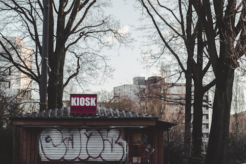 A small wooden kiosk with a corrugated metal roof stands in an urban setting. The kiosk has graffiti on its rolling door and a sign that reads 'KIOSK' on top. Surrounding the kiosk are bare trees with visible branches against a backdrop of high-rise buildings.