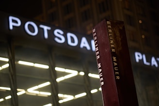 A metallic column with 'Berliner Mauer' written on it stands prominently, with large illuminated letters spelling 'POTSDAMER PLATZ' in the background. The scene appears to be set at night, with soft ambient lighting adding depth to the atmosphere.