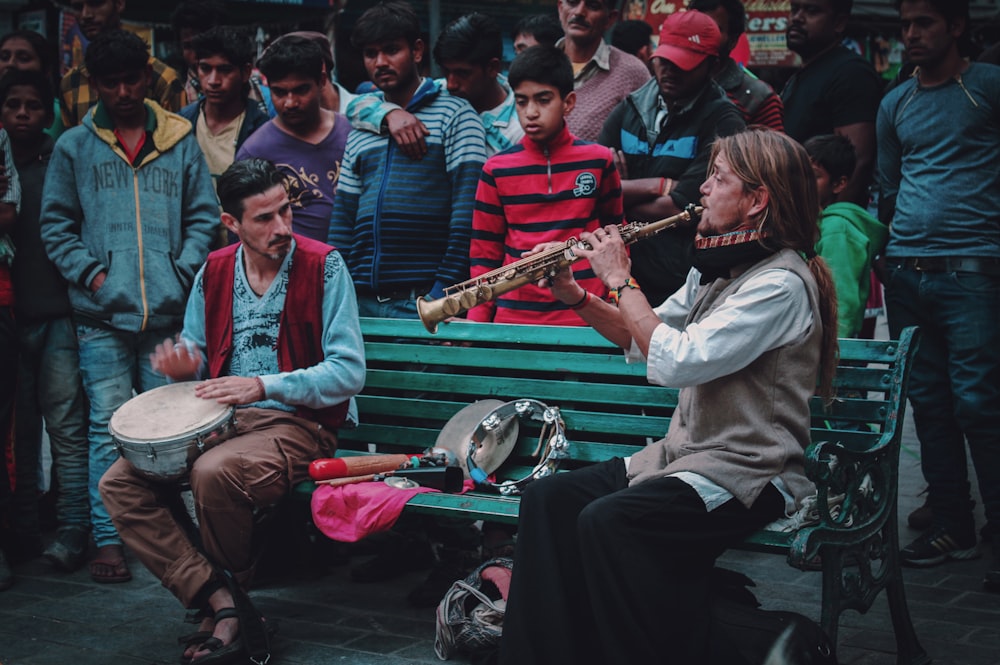 people gathering looking at the man sitting on bench playing wind instrument