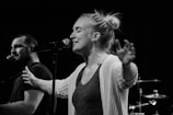 A black and white image featuring two people singing into microphones, likely performing on stage. The foreground shows a woman with arms extended, eyes closed, and engaged in singing. A man is visible in the background, also singing. Drum equipment is faintly visible in the background.