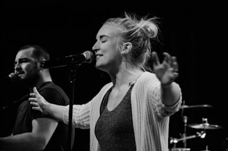 A black and white image featuring two people singing into microphones, likely performing on stage. The foreground shows a woman with arms extended, eyes closed, and engaged in singing. A man is visible in the background, also singing. Drum equipment is faintly visible in the background.