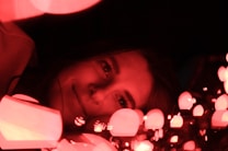 A person lying down surrounded by glowing red lights, creating a warm and intimate ambiance. The soft reflections from the lights illuminate the face, highlighting the eyes and a gentle smile.