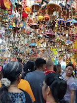 A crowded marketplace filled with vibrant and colorful hanging decorations, including bells, beads, and crafts. Many people are browsing through the lively and festive items displayed above and around them.