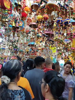 A crowded marketplace filled with vibrant and colorful hanging decorations, including bells, beads, and crafts. Many people are browsing through the lively and festive items displayed above and around them.
