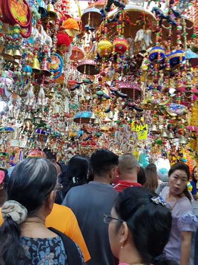 A crowded marketplace filled with vibrant and colorful hanging decorations, including bells, beads, and crafts. Many people are browsing through the lively and festive items displayed above and around them.