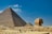 An expansive desert landscape features the iconic Great Pyramid and the Sphinx under a clear blue sky. Sparse groups of people are scattered around, providing a sense of scale to these ancient structures.