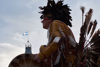 A person dressed in traditional Indigenous regalia, featuring feathers and intricate designs, appears to be participating in a cultural event. The background shows a clear sky with the Quebec flag flying and a building with a clock tower.