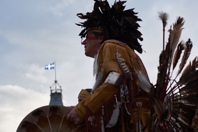 A person dressed in traditional Indigenous regalia, featuring feathers and intricate designs, appears to be participating in a cultural event. The background shows a clear sky with the Quebec flag flying and a building with a clock tower.