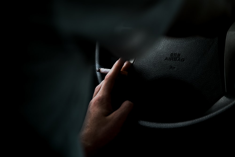 closeup photo of person's left hand on top of vehicle steering wheel while holding lighted cigarette