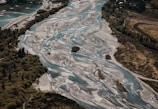 An aerial view of a braided river flowing through a landscape with lush green trees on the banks. The river has multiple channels and sandy areas, creating a striking pattern in contrast with surrounding vegetation. Hills and forested areas are visible, showcasing a natural and untouched environment.