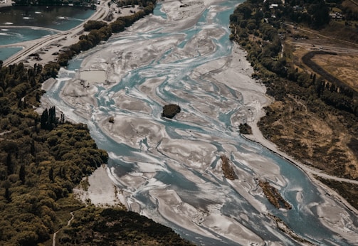 An aerial view of a braided river flowing through a landscape with lush green trees on the banks. The river has multiple channels and sandy areas, creating a striking pattern in contrast with surrounding vegetation. Hills and forested areas are visible, showcasing a natural and untouched environment.