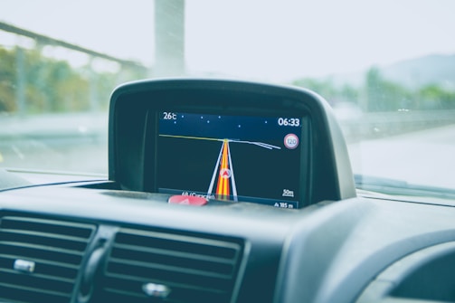 A car dashboard with a GPS navigation screen showing the current route. The screen displays various information including the temperature (26°C), time (06:33), and speed limit (120). The navigation path is highlighted with colored lines, and the remaining distance (50m) is also shown. Part of the car's air vents and windshield are visible.