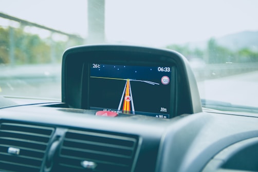 A car dashboard with a GPS navigation screen showing the current route. The screen displays various information including the temperature (26&deg;C), time (06:33), and speed limit (120). The navigation path is highlighted with colored lines, and the remaining distance (50m) is also shown. Part of the car