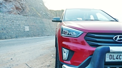 A red Hyundai car is parked on a road next to a rocky wall. The car's front view is prominently displayed, with visible headlights, grille, and a portion of the license plate.