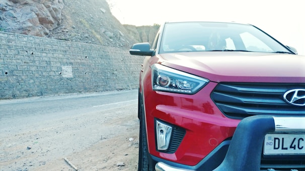 A red Hyundai car is parked on a road next to a rocky wall. The car's front view is prominently displayed, with visible headlights, grille, and a portion of the license plate.