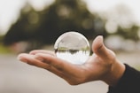 A hand is holding a clear crystal ball, which reflects an upside-down image of the surrounding environment. The background is blurred, focusing attention on the hand and the crystal ball.