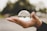 A hand is holding a clear crystal ball, which reflects an upside-down image of the surrounding environment. The background is blurred, focusing attention on the hand and the crystal ball.