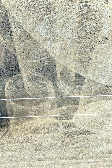 A textured concrete surface with subtle abstract patterns created by varying shades of gray. Two horizontal lines run across the image, possibly indicating cables or wires. Small granules add a sense of roughness and depth to the surface.