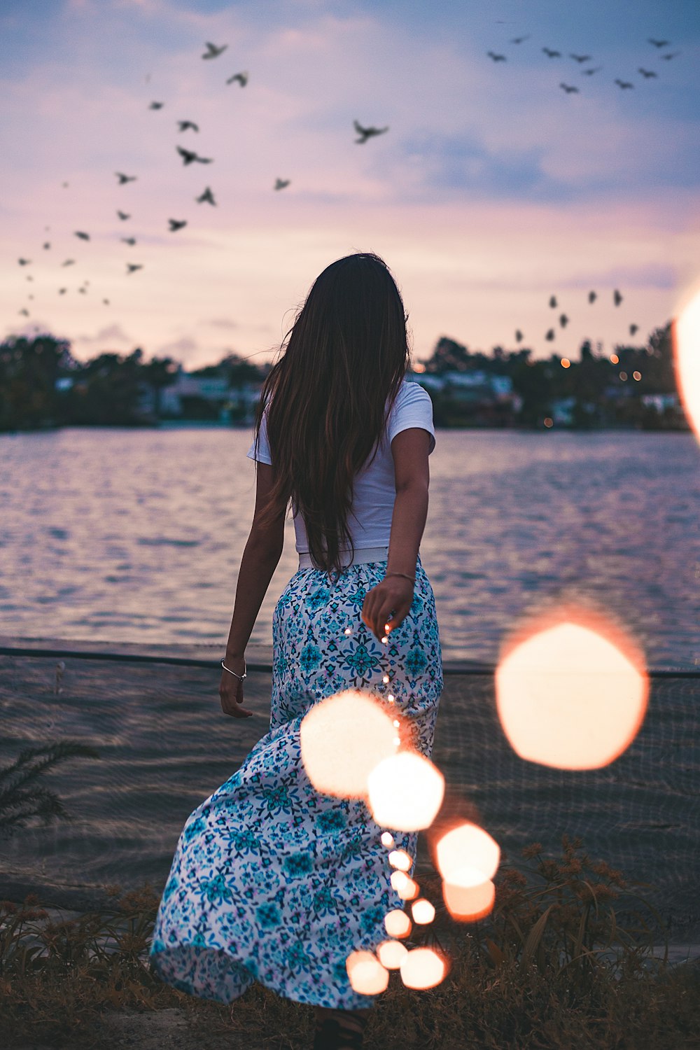 woman standing on green grass field in front body of water under flock of birds