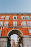 A vibrant orange building with white shutters and balconies features prominently, adorned with Spanish flags. An archway serves as a centerpiece, providing passage to a narrower street flanked by additional buildings. The sky above is clear and bright blue.