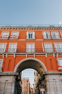 A vibrant orange building with white shutters and balconies features prominently, adorned with Spanish flags. An archway serves as a centerpiece, providing passage to a narrower street flanked by additional buildings. The sky above is clear and bright blue.