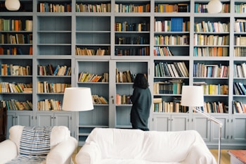 A person stands in front of a large bookshelf filled with a variety of books. The shelves are painted in a soft gray color and extend up to the ceiling. Two white couches are placed in the foreground alongside modern white lamps. The overall setting has a cozy and inviting atmosphere, ideal for reading and relaxation.