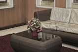 A wicker-style sofa with a glossy cushion is situated next to a matching coffee table. On the table, there's a decorative Christmas-themed floral arrangement in a red pot, featuring pine cones, greenery, and ornaments. The setting is completed by a maroon rug on a light-toned floor, and the background consists of wooden paneling and framed plaques.