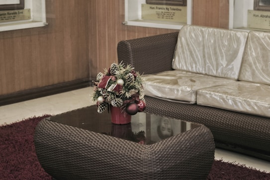 A wicker-style sofa with a glossy cushion is situated next to a matching coffee table. On the table, there's a decorative Christmas-themed floral arrangement in a red pot, featuring pine cones, greenery, and ornaments. The setting is completed by a maroon rug on a light-toned floor, and the background consists of wooden paneling and framed plaques.