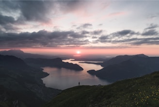silhouette of person standing on lake surrounded by mountains under cloudy sky during golden hour