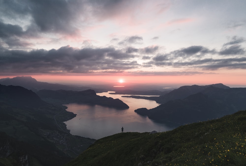 Silhouette einer Person, die während der goldenen Stunde auf dem See steht, umgeben von Bergen unter bewölktem Himmel