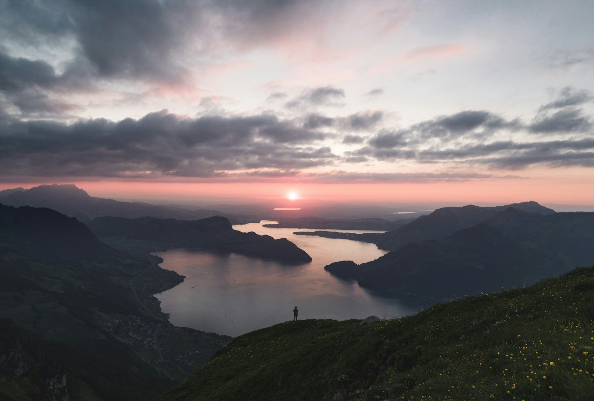 silhouette of person standing on lake surrounded by mountains under cloudy sky during golden hour