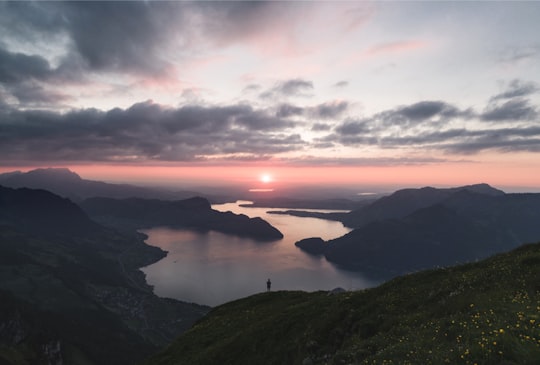 silhouette of person standing on lake surrounded by mountains under cloudy sky during golden hour in Niederbauen-Chulm Switzerland