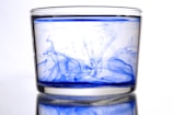 A clear glass filled with water is swirled with blue ink, creating vibrant patterns and swirls that spread through the liquid. The background is white, highlighting the contrast of the blue ink against the transparent water.