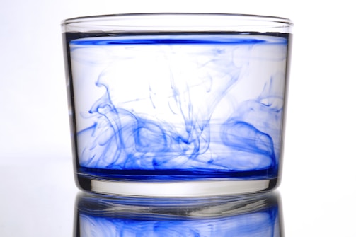 A clear glass filled with water is swirled with blue ink, creating vibrant patterns and swirls that spread through the liquid. The background is white, highlighting the contrast of the blue ink against the transparent water.