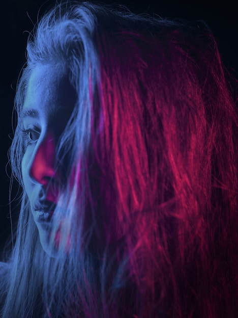 A woman's profile is captured with vibrant lighting. Her face is partially illuminated with blue and pink light, creating a striking contrast against the dark background. The colors blend into her long hair, giving a dramatic effect.